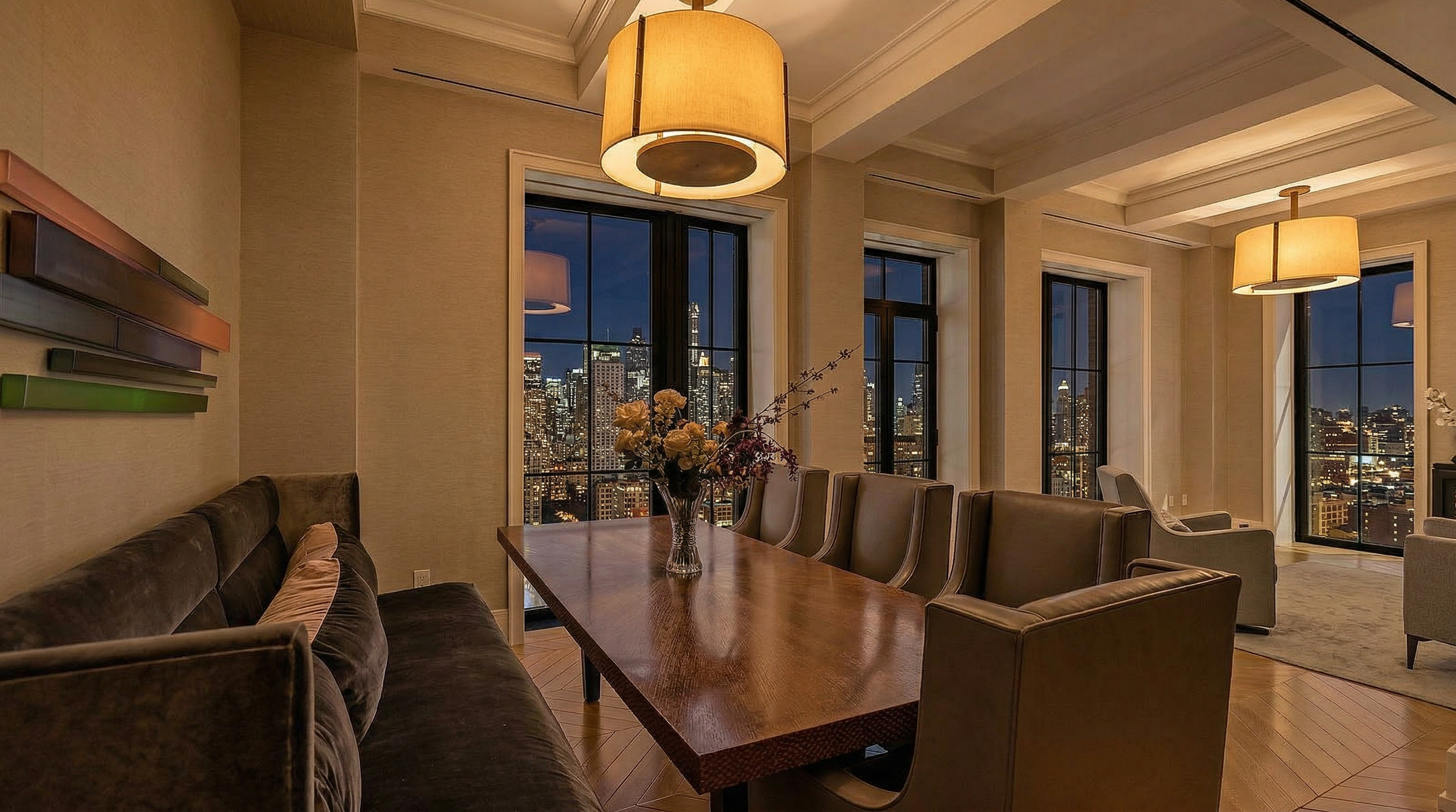 This elegant dining room features a long, dark-finished wooden table paired with sophisticated gray upholstered chairs and a velvet banquet seating area. Large floor-to-ceiling windows offer a breathtaking nocturnal view of the city skyline, complemented by warm pendant lighting that creates an inviting atmosphere. The space blends contemporary luxury with a refined design aesthetic, perfect for formal entertaining.