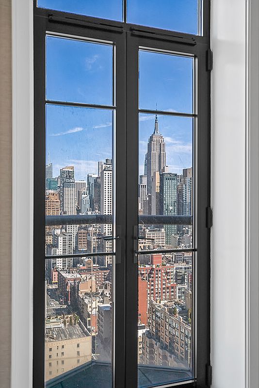 This image captures a tall, black-framed French door or window set against a clean white wall, framing a stunning, high-altitude view of the New York City skyline. The central focus is the iconic Empire State Building, visible through the clear glass panes, emphasizing the property's prime urban location and dramatic perspective. The sleek, minimalist design of the window frame provides a sophisticated contrast to the dense, bustling cityscape beyond.