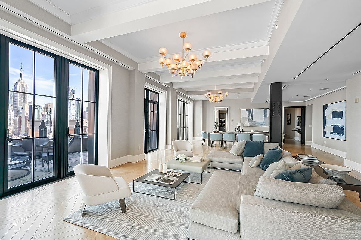 This expansive, high-end living room features a sophisticated open-concept layout with light hardwood floors in a herringbone pattern and elegant coffered ceilings. The space is anchored by a large, plush sectional sofa and a modern coffee table, while floor-to-ceiling windows provide a stunning, iconic view of the New York City skyline. A formal dining area is visible in the background, illuminated by a matching contemporary chandelier, creating a seamless flow perfect for luxury entertaining.