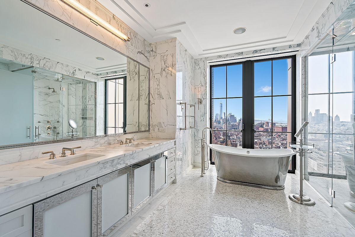 This luxurious primary bathroom features high-end marble finishes, a double vanity with integrated sinks, and a sleek, metallic soaking tub positioned to take advantage of the expansive city skyline view. The room is bathed in natural light from floor-to-ceiling windows, creating an airy and sophisticated atmosphere. A glass-enclosed shower and polished stone flooring complete the elegant, modern design.