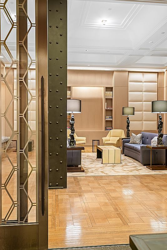 This sophisticated lobby or lounge area features a refined, high-end aesthetic with a blend of Art Deco-inspired metalwork and modern luxury furnishings. The space is anchored by a polished parquet wood floor, plush seating, and unique sculptural table lamps, all set against a backdrop of elegant, paneled walls and built-in shelving. The perspective is framed through a decorative metal screen, creating a sense of exclusivity and architectural depth.