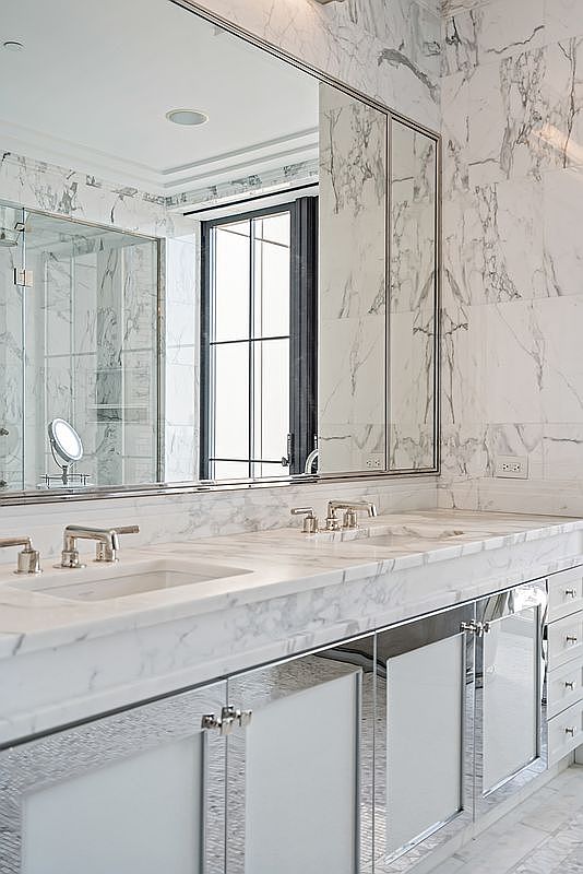 This luxurious primary bathroom features a double vanity with a polished marble countertop and integrated sinks, complemented by a large, seamless mirror that reflects the elegant marble-tiled walls. The cabinetry below is finished with mirrored panels and chrome hardware, creating a sophisticated and bright aesthetic. A large window with a dark frame adds a modern architectural contrast to the classic stone surfaces.