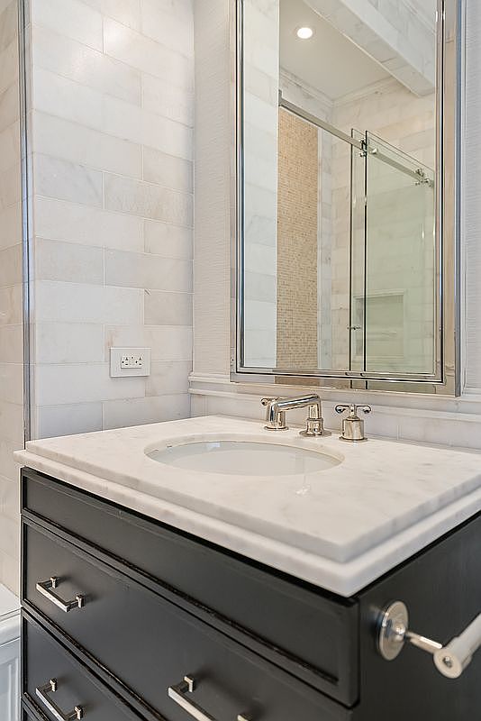 This elegant bathroom vanity features a dark charcoal cabinet topped with a white marble countertop and an undermount sink. A polished chrome faucet and matching hardware complement the sophisticated aesthetic, while the wall is finished with classic white subway tiles. A large, framed mirror reflects the shower area, creating a bright and polished look suitable for a high-end home.