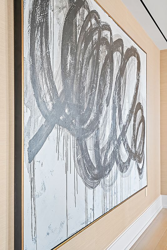 This image captures a close-up, angled perspective of a large-scale abstract painting mounted on a wall covered in textured, neutral-toned wallpaper. The artwork features bold, dark gray gestural brushstrokes and drips against a light, distressed background, framed in a sleek, dark-edged frame with gold accents. The composition emphasizes the artistic focal point of a sophisticated interior space, highlighting the high-end finishes and modern aesthetic.