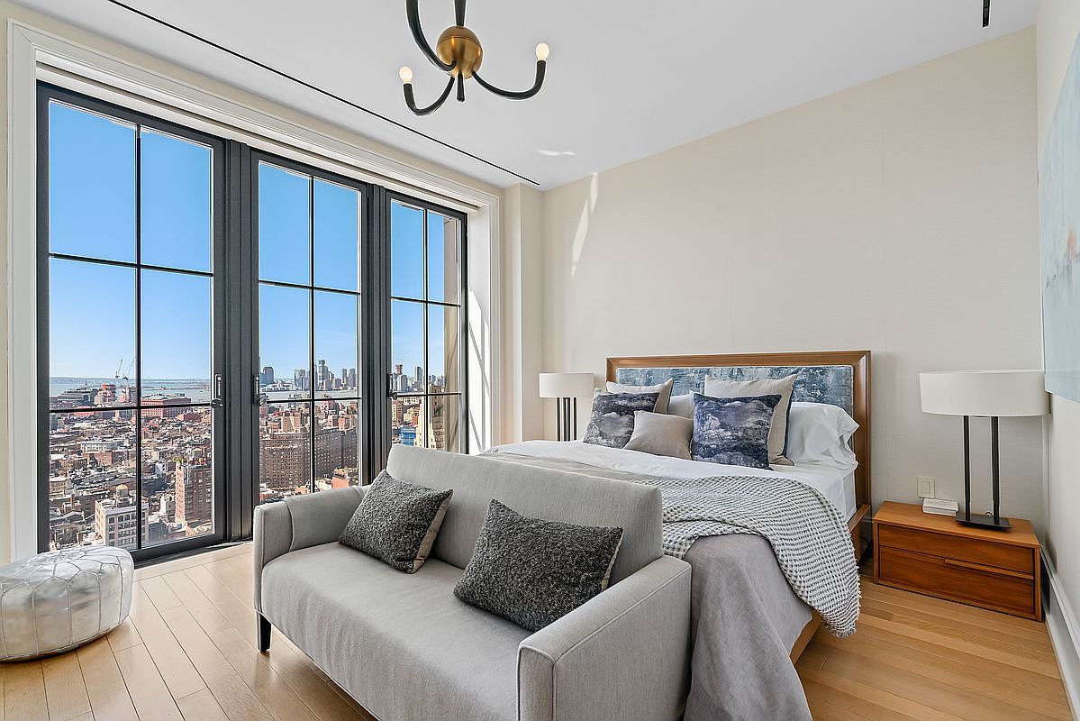 This sophisticated primary bedroom features a contemporary design with a plush bed, a sleek gray sofa at its foot, and a striking modern chandelier. Large floor-to-ceiling windows provide an expansive, breathtaking view of the city skyline and harbor, bathing the room in natural light. The neutral color palette and clean lines create a serene, high-end urban retreat.
