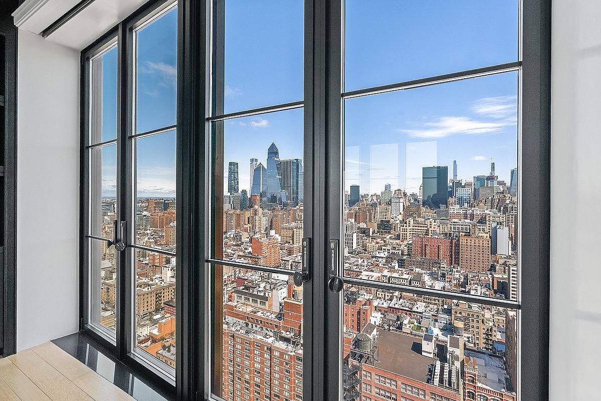 This interior shot captures a stunning, high-rise perspective of a dense city skyline through floor-to-ceiling black-framed casement windows. The minimalist design emphasizes the expansive urban view, highlighting the luxury of height and natural light. The clean lines of the window frames and the light-colored flooring suggest a modern, sophisticated living space.