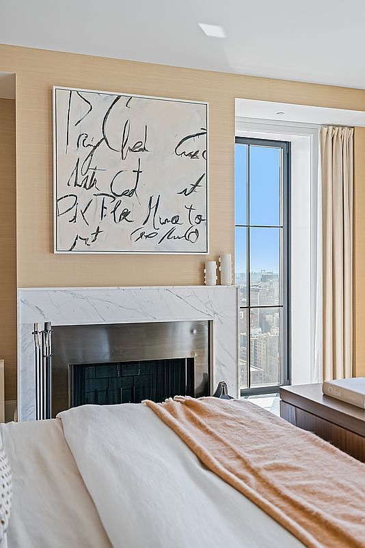 This elegant bedroom features a sophisticated fireplace with a white marble surround and a sleek metallic firebox, topped by a large, abstract calligraphic art piece. A plush bed with a soft peach-toned throw blanket occupies the foreground, while a tall window to the right offers a stunning, high-rise urban view. The room is finished with textured, neutral-toned wallpaper, creating a serene and luxurious atmosphere.