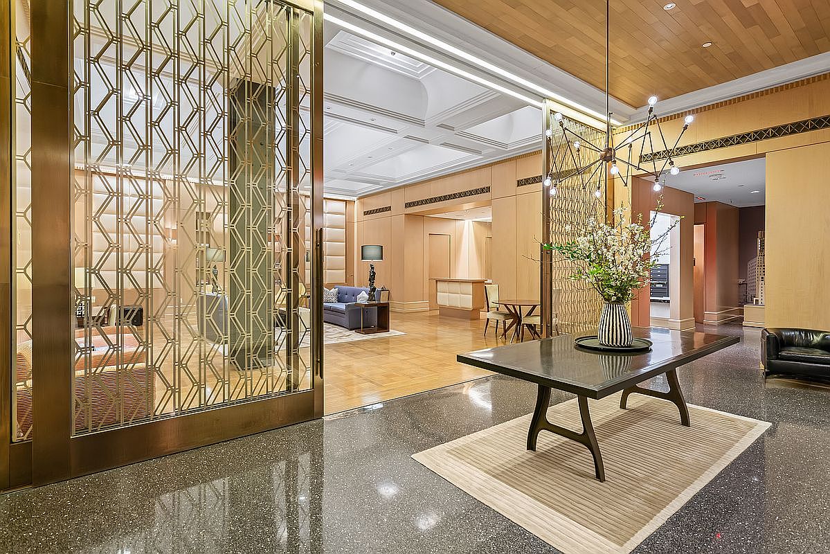 This sophisticated lobby or common area features a high-end, modern aesthetic with a striking gold-toned geometric metal screen partition. A sleek dark wood console table topped with a floral arrangement sits on a textured rug, while the polished terrazzo flooring reflects the warm, ambient lighting. The space conveys a sense of luxury and exclusivity, typical of a high-end residential building or hotel entrance.