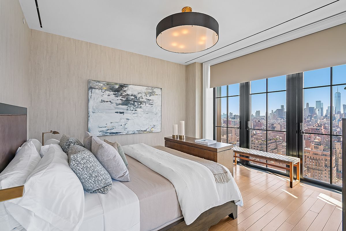This sophisticated primary bedroom features a plush king-sized bed with layered neutral bedding, positioned to take full advantage of the floor-to-ceiling windows that offer a breathtaking panoramic view of the city skyline. The room is accented by a large abstract painting, a sleek wooden console table, and a modern drum-style chandelier, creating an atmosphere of refined urban luxury. The warm wood flooring and soft, textured wall coverings enhance the serene and upscale aesthetic of this high-rise retreat.