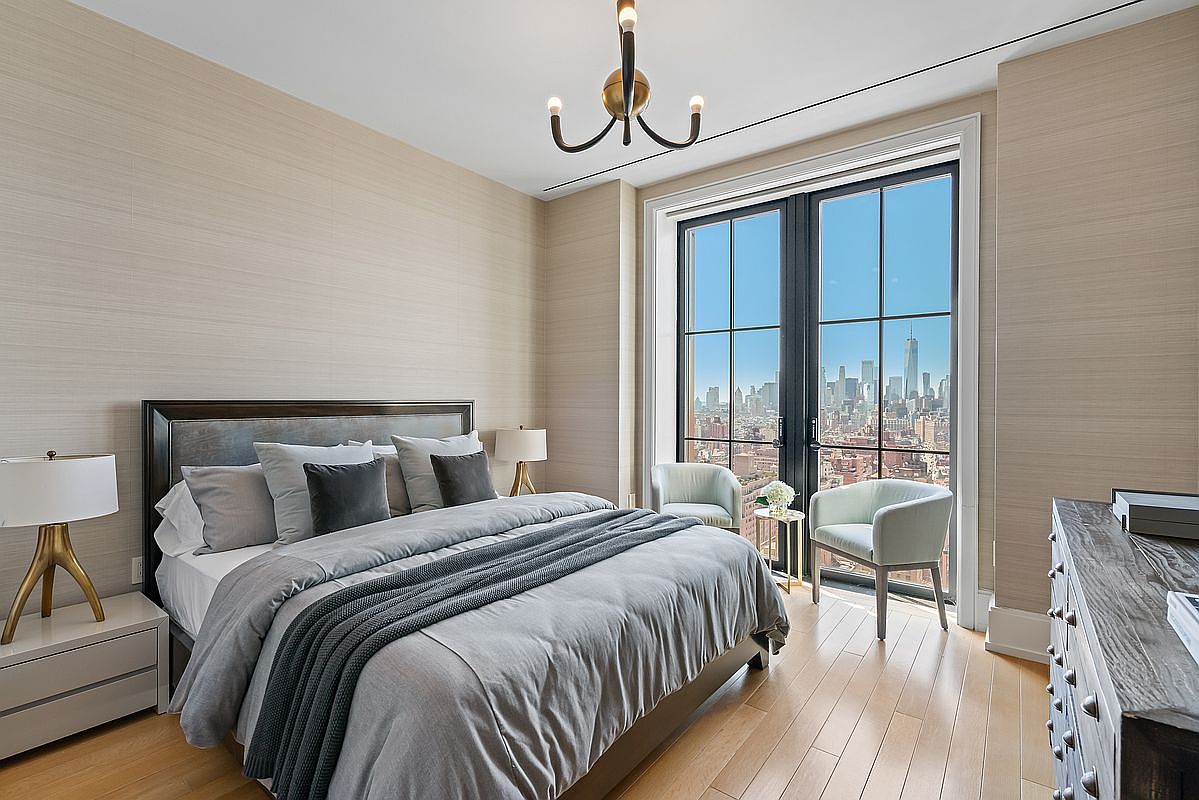 This sophisticated primary bedroom features a neutral color palette with textured wallpaper and a plush, modern bed dressed in layered gray linens. A large floor-to-ceiling window provides a stunning, unobstructed view of the city skyline, while two elegant armchairs create a cozy seating area. The room is illuminated by a contemporary chandelier, blending luxury with a serene, high-rise urban atmosphere.