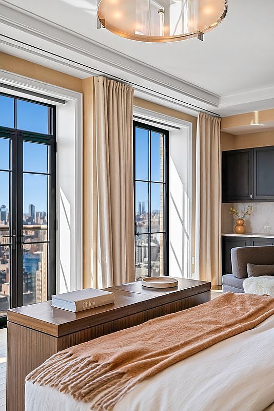 This elegant primary bedroom features a sophisticated, neutral color palette with a plush bed topped by a textured, warm-toned throw blanket. Large, floor-to-ceiling windows with floor-length curtains frame a stunning, expansive city skyline view, while a modern, circular chandelier adds a touch of luxury. The space is anchored by a sleek wooden console at the foot of the bed, creating a refined and tranquil atmosphere.
