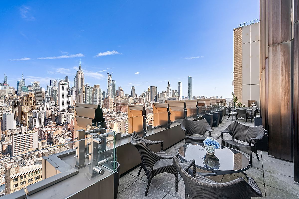 This high-rise balcony offers a sophisticated outdoor living space with a panoramic view of the New York City skyline, including the Empire State Building. The area is furnished with modern, dark-wicker patio furniture arranged around a glass-topped table, set against a backdrop of sleek metal and glass railings. The perspective captures the vast urban landscape from an elevated vantage point, emphasizing the luxury and exclusivity of the property.