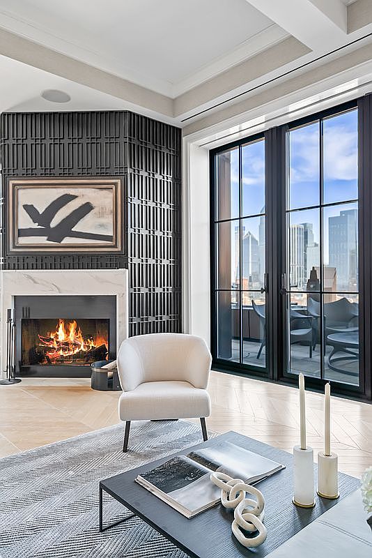This sophisticated living room features a striking black slatted accent wall surrounding a marble-framed fireplace with a roaring fire. A plush, cream-colored armchair sits atop a textured gray rug, while a sleek black coffee table holds decorative accents and candles. Large floor-to-ceiling windows provide a stunning, expansive view of the city skyline, creating a luxurious and serene urban atmosphere.