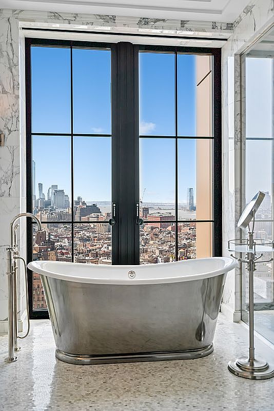 This luxurious primary bathroom features a sleek, metallic freestanding soaking tub positioned directly in front of a floor-to-ceiling window that offers a breathtaking, panoramic view of the city skyline. The space is finished with elegant marble wall cladding and a textured, light-colored tiled floor, complemented by a modern chrome floor-mounted faucet and a matching pedestal vanity mirror. The overall aesthetic is one of sophisticated urban opulence, emphasizing high-end materials and a dramatic, light-filled atmosphere.