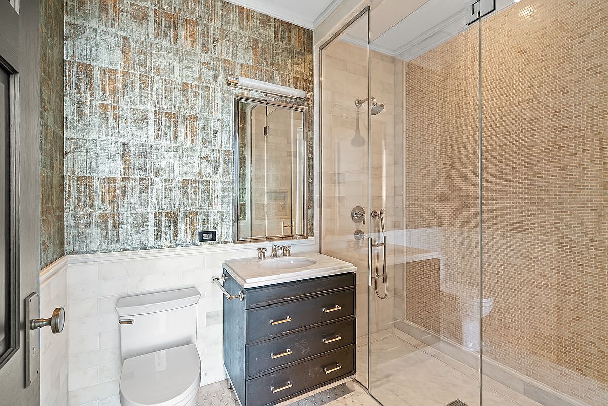 This elegant bathroom features a sophisticated blend of textures, including a metallic-finish wallpaper above white marble wainscoting and a glass-enclosed walk-in shower with mosaic tile accents. A dark, multi-drawer vanity with a marble countertop and a sleek, framed mirror adds a touch of modern luxury to the space. The perspective is a wide-angle shot that captures the entire layout, emphasizing the high-end finishes and clean, contemporary design.