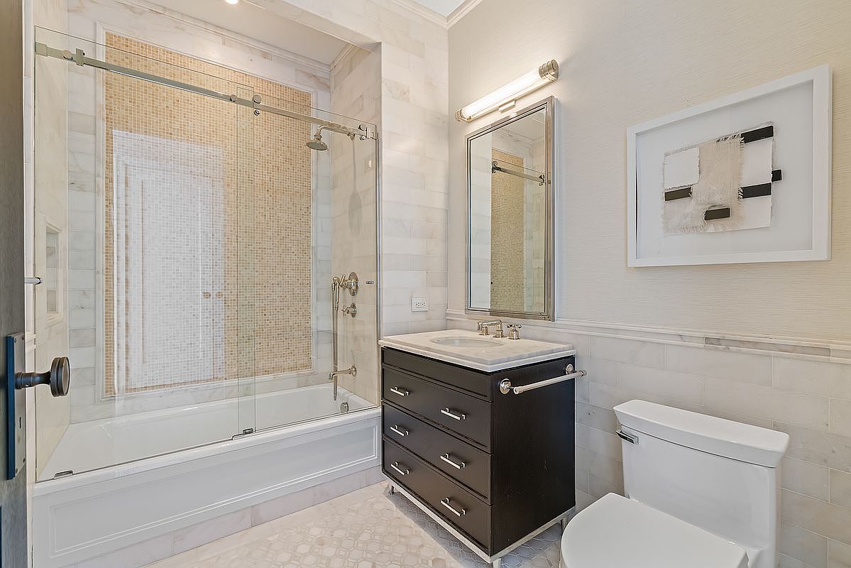 This elegant bathroom features a sleek, dark wood vanity with a marble countertop and a modern, rectangular undermount sink. A glass-enclosed shower-tub combination is accented with a textured, neutral-toned mosaic tile wall, while the rest of the room is finished in classic white subway tile. The space is brightened by a contemporary vanity light above a framed mirror and a piece of abstract wall art, creating a sophisticated and clean aesthetic.