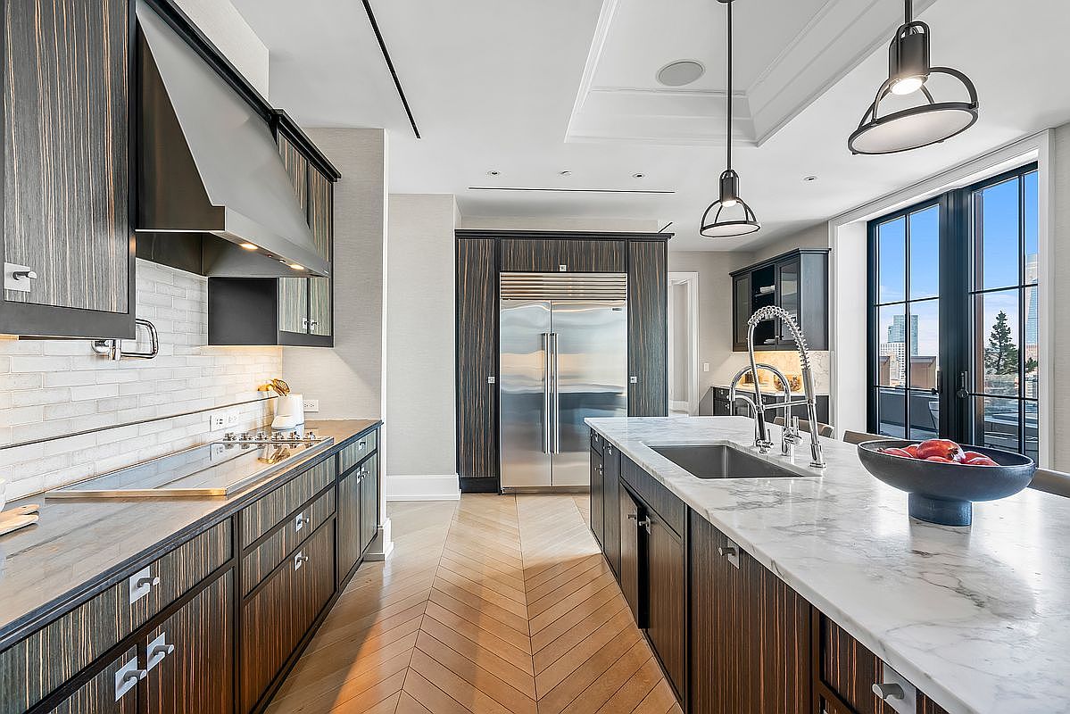 This sophisticated, high-end kitchen features dark, vertically-grained wood cabinetry paired with sleek marble countertops and a matching backsplash. A large central island with a stainless steel sink serves as a focal point, complemented by modern pendant lighting and a professional-grade stainless steel refrigerator. The space is anchored by light-toned herringbone wood flooring, creating a warm contrast against the dark cabinetry and offering a bright, airy atmosphere with views through the adjacent balcony doors.