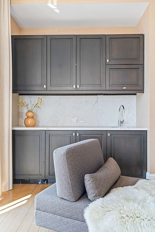This image features a sophisticated wet bar or kitchenette area characterized by dark, matte-finished cabinetry and a crisp white marble backsplash and countertop. A modern chrome faucet is integrated into the sink, while a decorative vase adds a touch of warmth to the sleek, minimalist design. In the foreground, a plush, textured gray lounge chair with a faux fur throw creates a cozy, inviting atmosphere, suggesting a seamless blend of functional utility and relaxed living space.