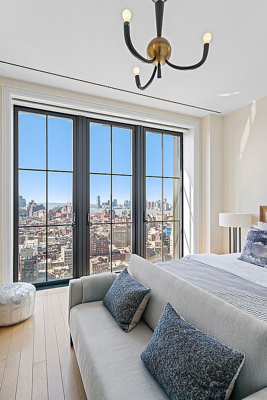 This sophisticated bedroom features a light-filled, modern aesthetic with a plush sofa positioned at the foot of the bed, creating a cozy seating area. Large, floor-to-ceiling black-framed windows offer a breathtaking, expansive view of the city skyline. A contemporary chandelier hangs from the ceiling, adding a touch of elegance to the minimalist, high-end design.