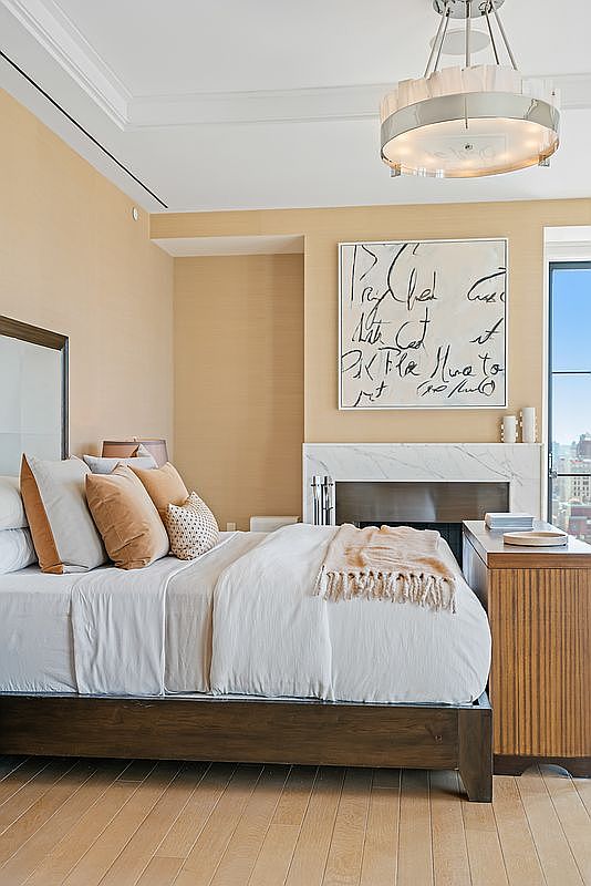 This elegant primary bedroom features a sophisticated neutral color palette, highlighted by textured beige wallpaper and a warm wooden bed frame. A marble-surround fireplace serves as a focal point, topped with abstract art, while a modern tiered chandelier adds a touch of luxury. The room is bathed in natural light from a large window, creating a serene and high-end atmosphere.