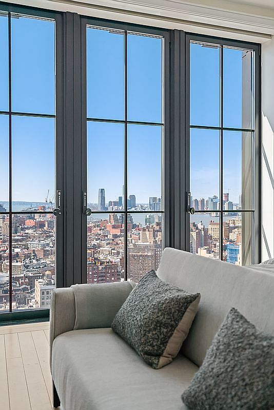 This interior shot captures a sophisticated living room space featuring a plush, light-colored sofa adorned with textured grey throw pillows. The focal point is a striking floor-to-ceiling window wall with dark frames, offering a breathtaking, expansive panoramic view of a dense city skyline and waterfront. The perspective is intimate and inviting, emphasizing the seamless blend of luxurious indoor comfort and the dramatic urban landscape beyond.