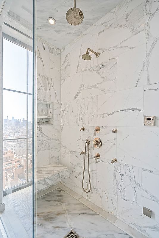 This luxurious walk-in shower features floor-to-ceiling white marble tiling with dramatic grey veining, creating a sophisticated and high-end aesthetic. The space includes a built-in marble bench, a rainfall showerhead, a handheld sprayer, and a wall-mounted digital control panel, all finished in polished gold. A large floor-to-ceiling window provides a stunning, expansive view of the city skyline, blending modern urban living with spa-like tranquility.
