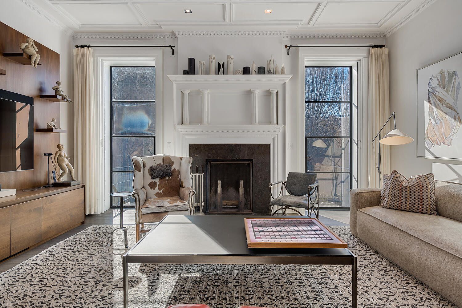 This is a well-appointed living room featuring a fireplace with decorative mantel, flanked by large windows with elegant drapery. The room is furnished with a sofa, armchairs, and a coffee table with a game board, all set on a patterned rug. The perspective is from the center of the room, capturing the entire space and highlighting the architectural details and sophisticated decor.