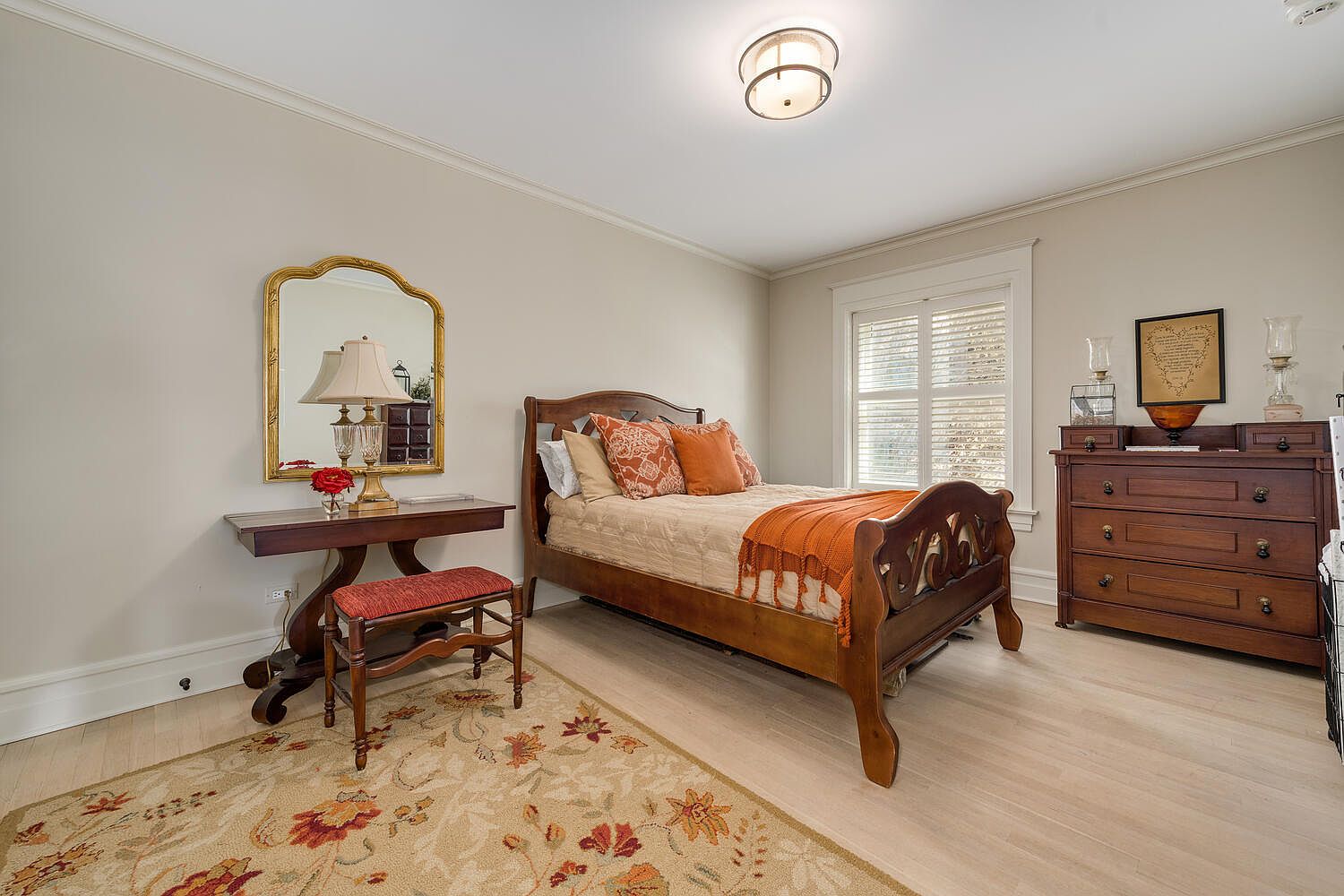 This is a cozy guest bedroom featuring a wooden bed frame with orange and patterned pillows, complemented by a matching throw blanket. A small wooden desk with a gold-framed mirror and a cushioned stool sit to the side, while a wooden dresser stands against the wall near the window. The room is decorated with a floral rug and a classic ceiling light fixture, creating a warm and inviting atmosphere.
