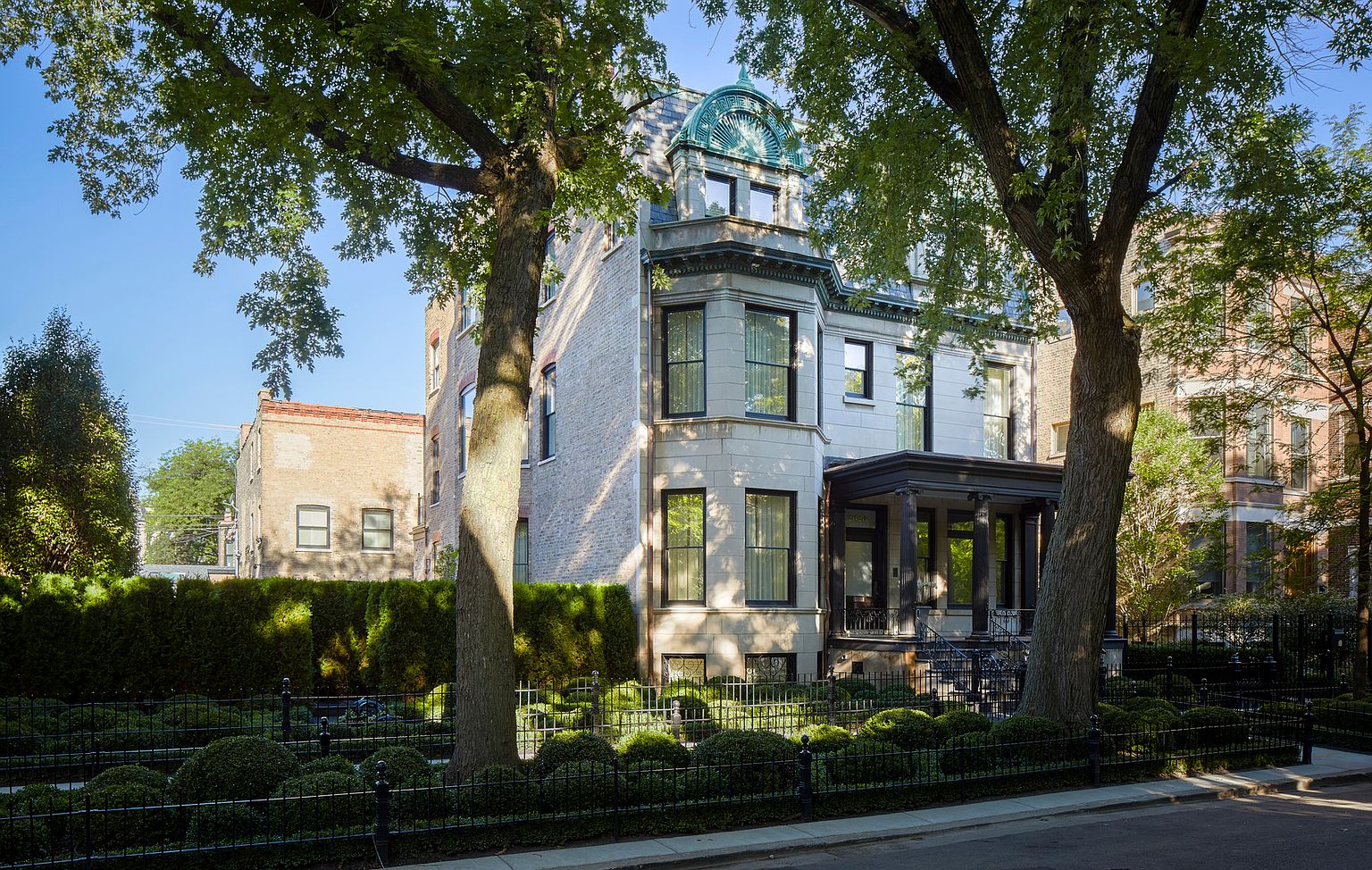 This is a front exterior view of a stately multi-story home with a classic architectural style. The facade features a combination of brick and stone, with large windows and a covered porch supported by columns. The property is surrounded by a well-maintained garden with a wrought-iron fence, enhancing its curb appeal.
