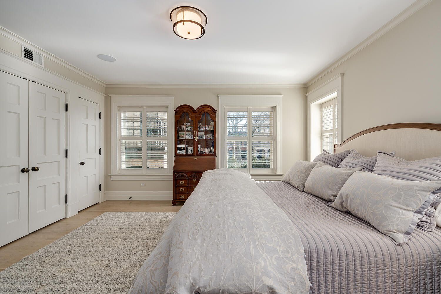 This is a well-lit primary bedroom featuring a large bed with a padded headboard and multiple pillows, a light-colored rug, and built-in closets. Two windows with blinds flank a wooden secretary desk, adding character to the room. The overall style is traditional and comfortable, creating a relaxing atmosphere.