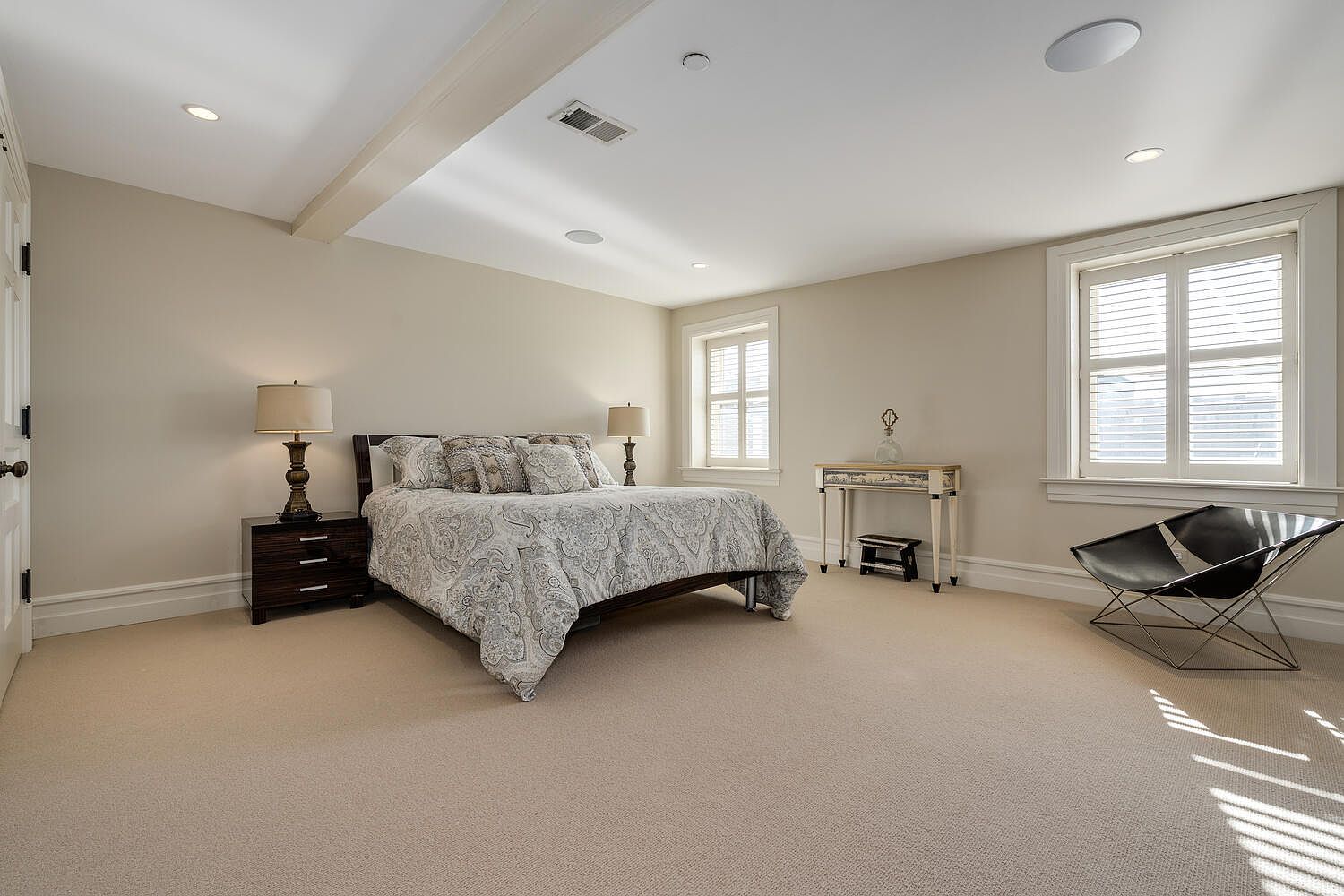 This is a well-lit primary bedroom featuring neutral-toned walls and carpet. The room includes a bed with patterned bedding, two bedside tables with lamps, and a small desk with a chair. Natural light streams in through shuttered windows, creating a calm and inviting atmosphere.