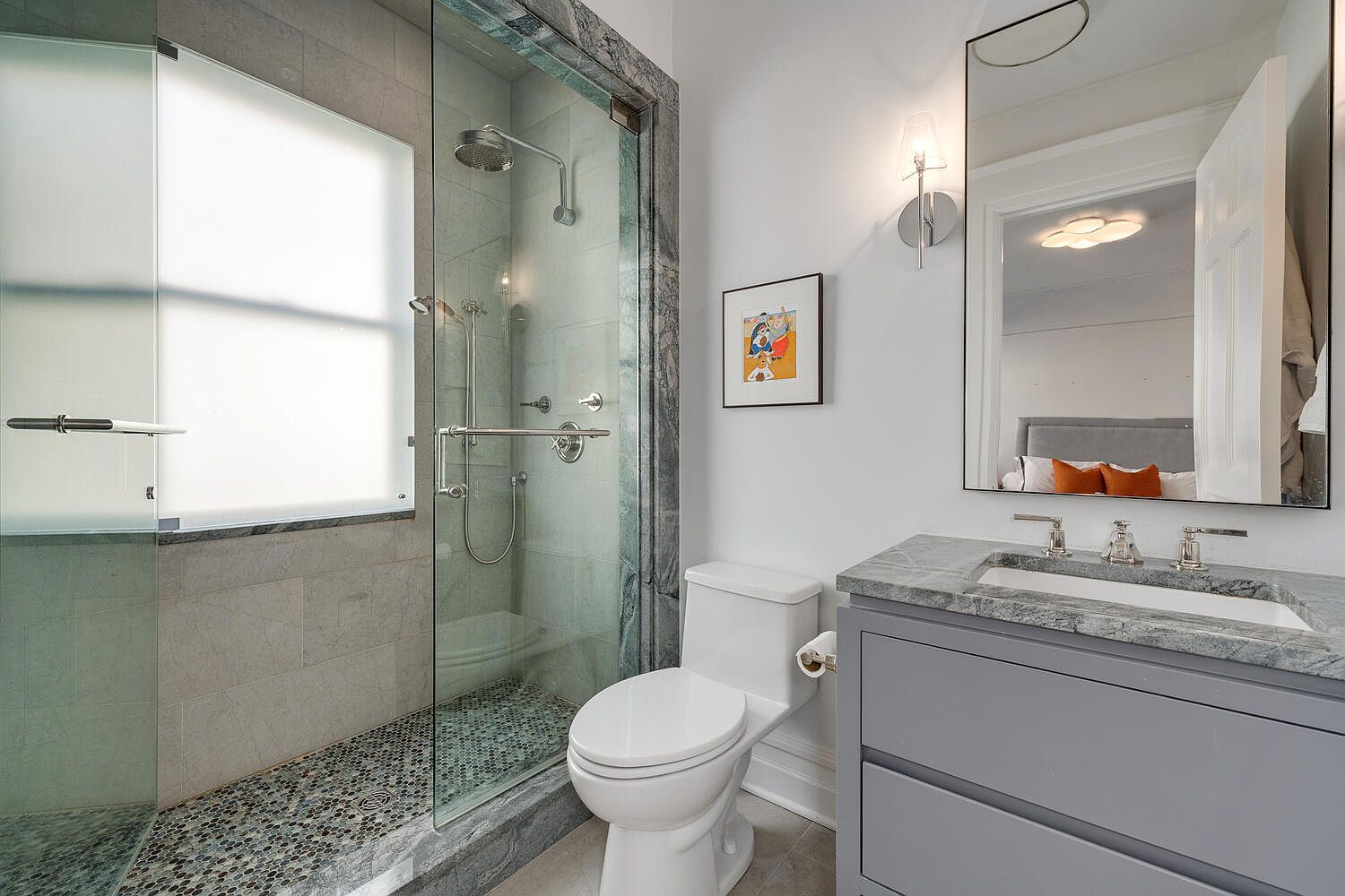 This is a well-lit bathroom featuring a glass-enclosed shower with mosaic tile flooring and a frosted window. A toilet sits next to the shower, and a gray vanity with a marble countertop and a large mirror completes the space. The room has a clean and modern aesthetic.
