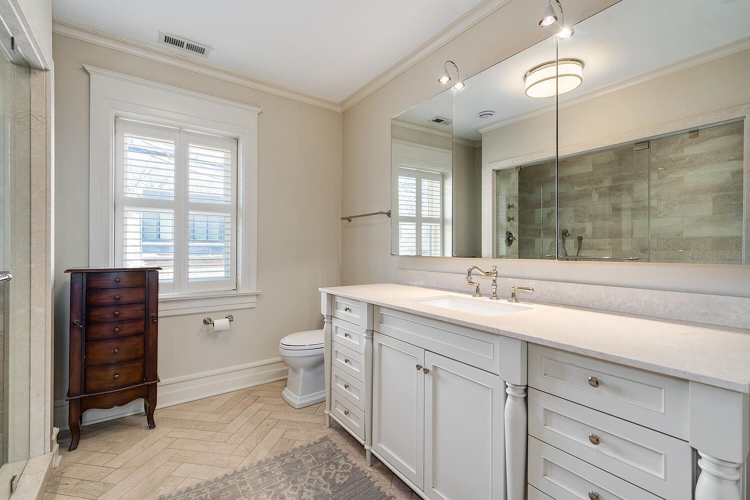This is a well-lit primary bathroom featuring a large mirror above a white vanity with ample storage. A toilet is visible to the left of the vanity, and a dark wood cabinet stands near a window with white shutters. The floor is tiled in a herringbone pattern, and a gray rug adds a touch of comfort.