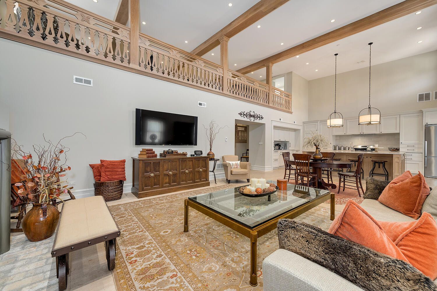 This is an interior shot of a spacious living room with a high ceiling and a second-floor balcony overlooking the area. The room features a large TV, a wooden cabinet, a glass-topped coffee table, and comfortable seating. The overall style is traditional with modern touches, creating a warm and inviting atmosphere.