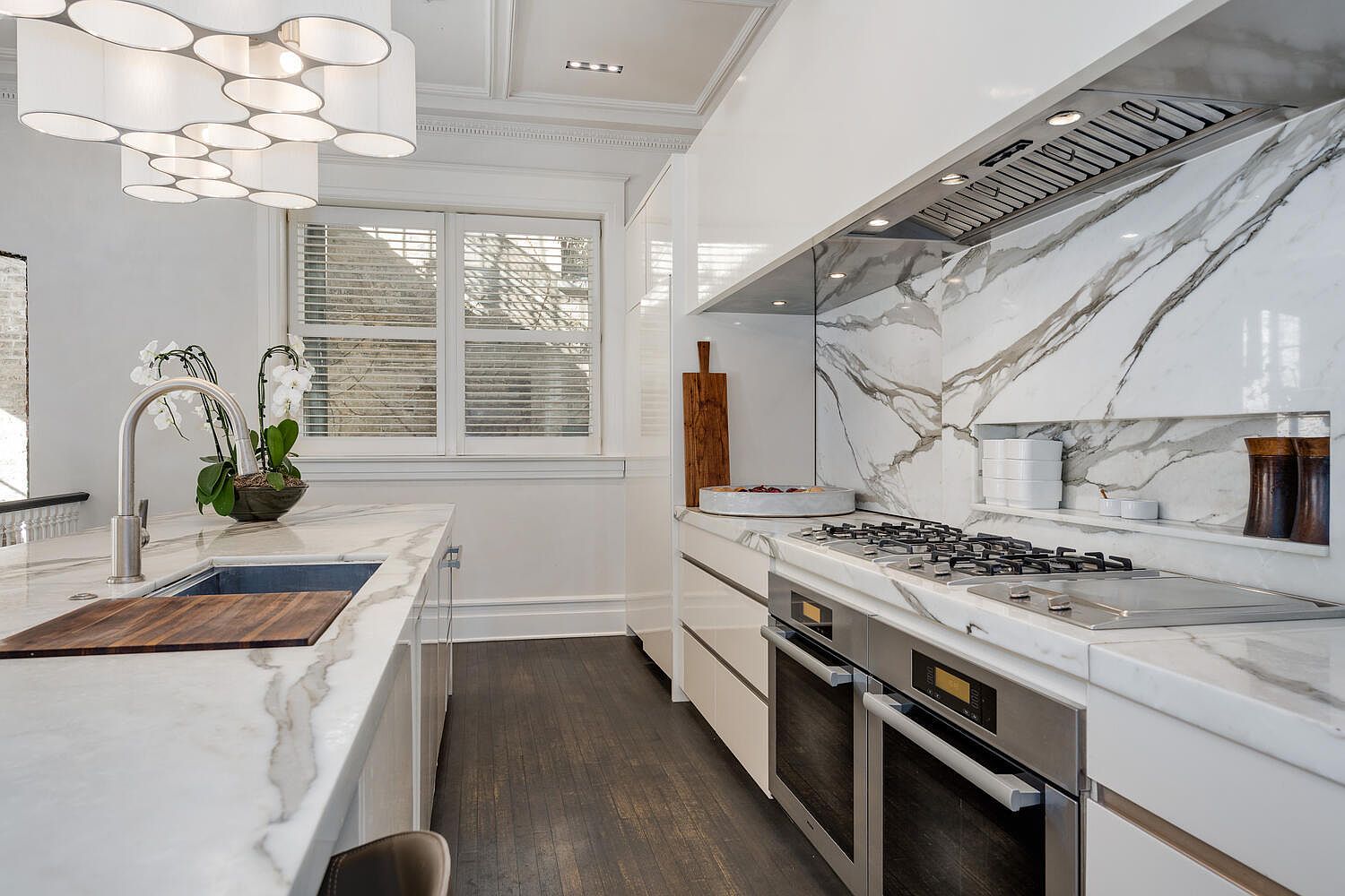 This is a bright, modern kitchen featuring white marble countertops and backsplash with striking gray veining. Stainless steel appliances, including double ovens and a gas cooktop, are seamlessly integrated into the cabinetry. A large island with a sink and wooden cutting board provides ample workspace, and a unique, multi-bulb chandelier illuminates the space.