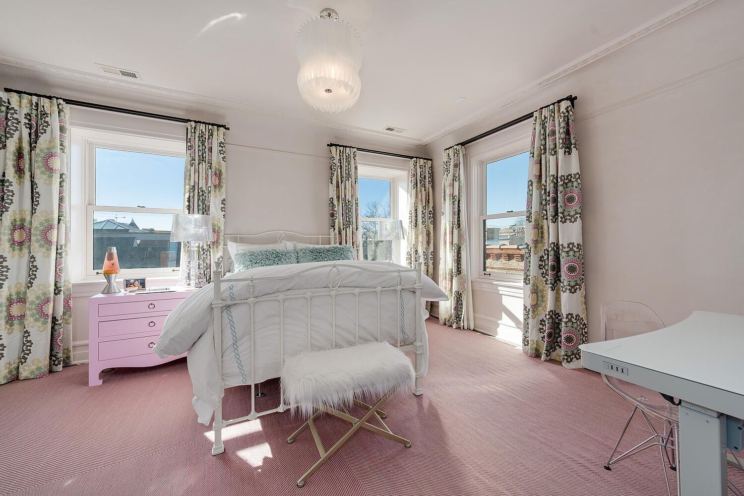 This is a bright and airy bedroom featuring a white iron bed frame, a pink dresser, and patterned curtains that frame the windows. The room is decorated in a whimsical style, with a fluffy white bench at the foot of the bed and a modern desk and chair in the corner. The pink herringbone carpet adds a touch of warmth and texture to the space.