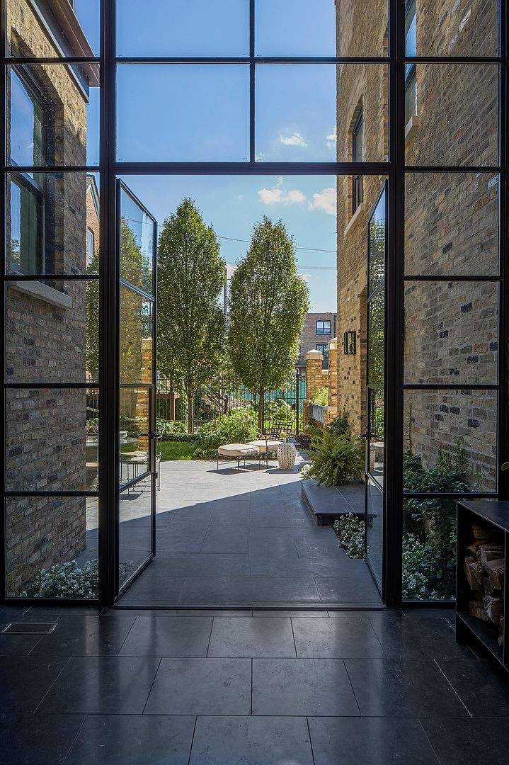 This image showcases a beautifully landscaped backyard viewed through large, modern glass doors. The patio features sleek gray tiling, leading to a well-maintained garden with mature trees and stylish outdoor furniture. The brick exterior of the building adds a touch of classic charm, creating an inviting and sophisticated outdoor living space.