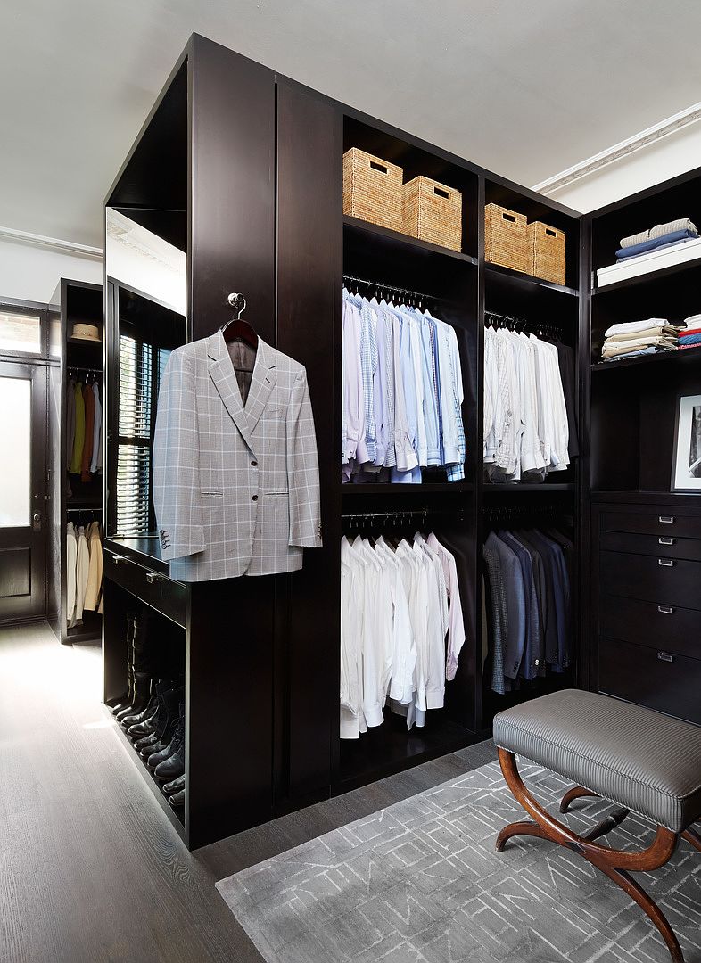 This is a well-organized walk-in closet featuring dark wood cabinetry and ample storage space. The closet includes hanging rods for shirts and suits, shelves for folded clothes and woven baskets, and a dedicated area for shoes. A gray patterned rug and a cushioned bench add a touch of luxury to the space.