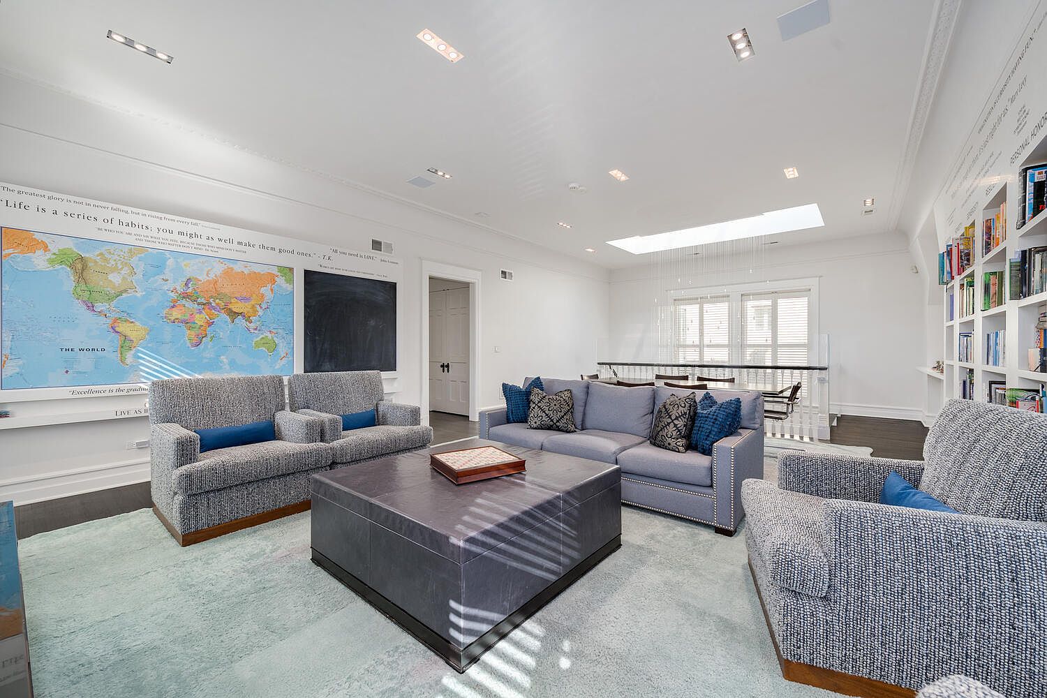 This is a spacious and well-lit living room featuring a world map and chalkboard on one wall, complemented by inspirational quotes. The room is furnished with a light blue sofa, two armchairs, and a large square coffee table, all set on a light blue rug. Natural light floods the space through a skylight, enhancing the bright and airy atmosphere.