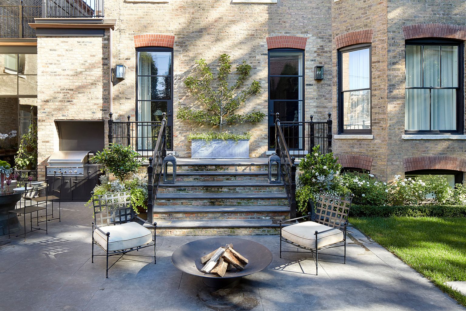 This is a beautifully designed outdoor patio area, featuring a fire pit surrounded by comfortable seating. The patio is accessed by stone steps leading up to a brick building with elegant windows and wrought iron railings. A built-in grill area is visible on the left, suggesting an outdoor kitchen setup, and the landscaping includes manicured grass and flowering plants, creating a serene and inviting atmosphere.