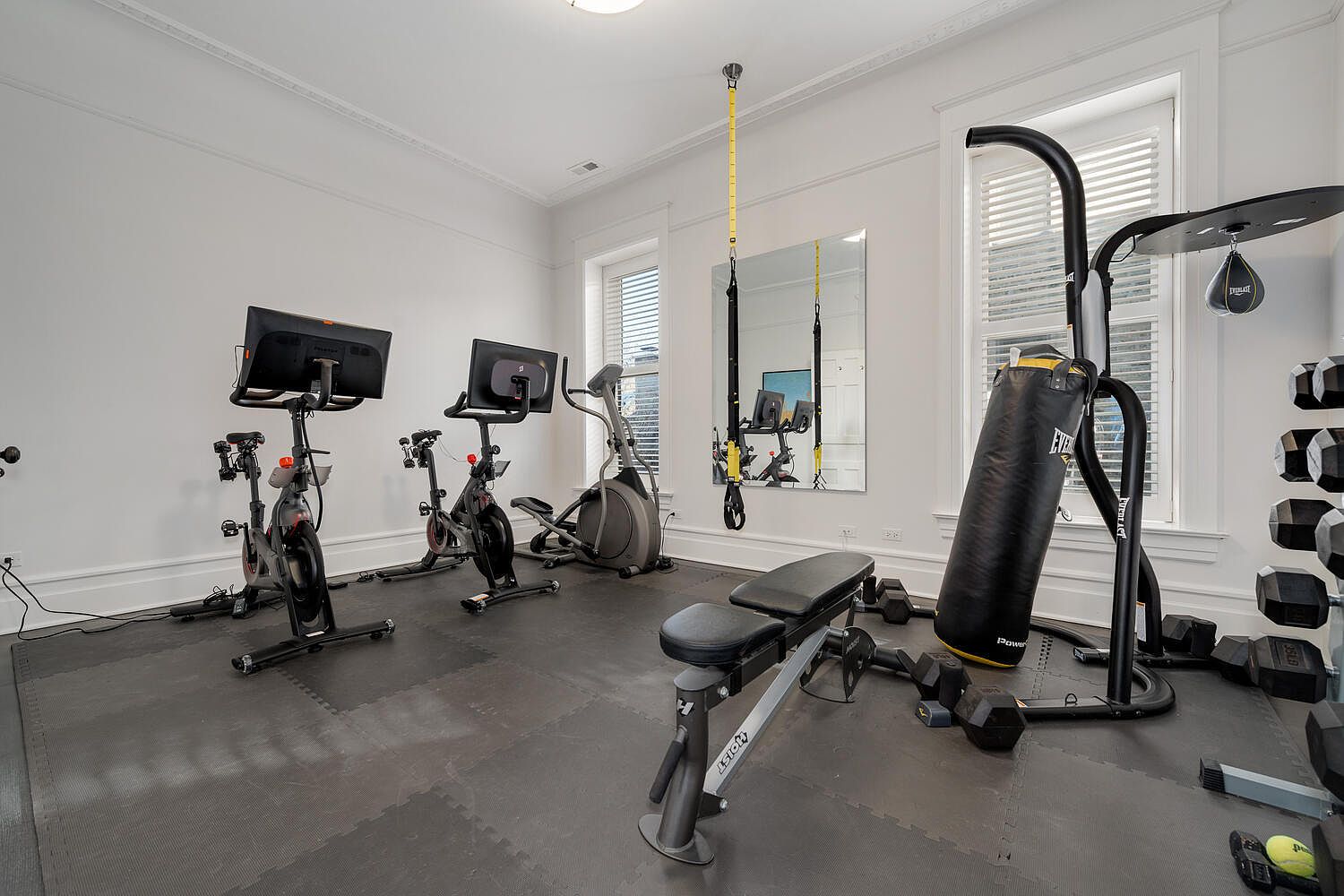 This is a well-equipped home gym featuring two Peloton bikes, an elliptical machine, a weight bench, a punching bag, and a dumbbell rack. The room has a black rubber floor, white walls, and natural light coming through the windows. The overall impression is a clean and functional space for exercise.