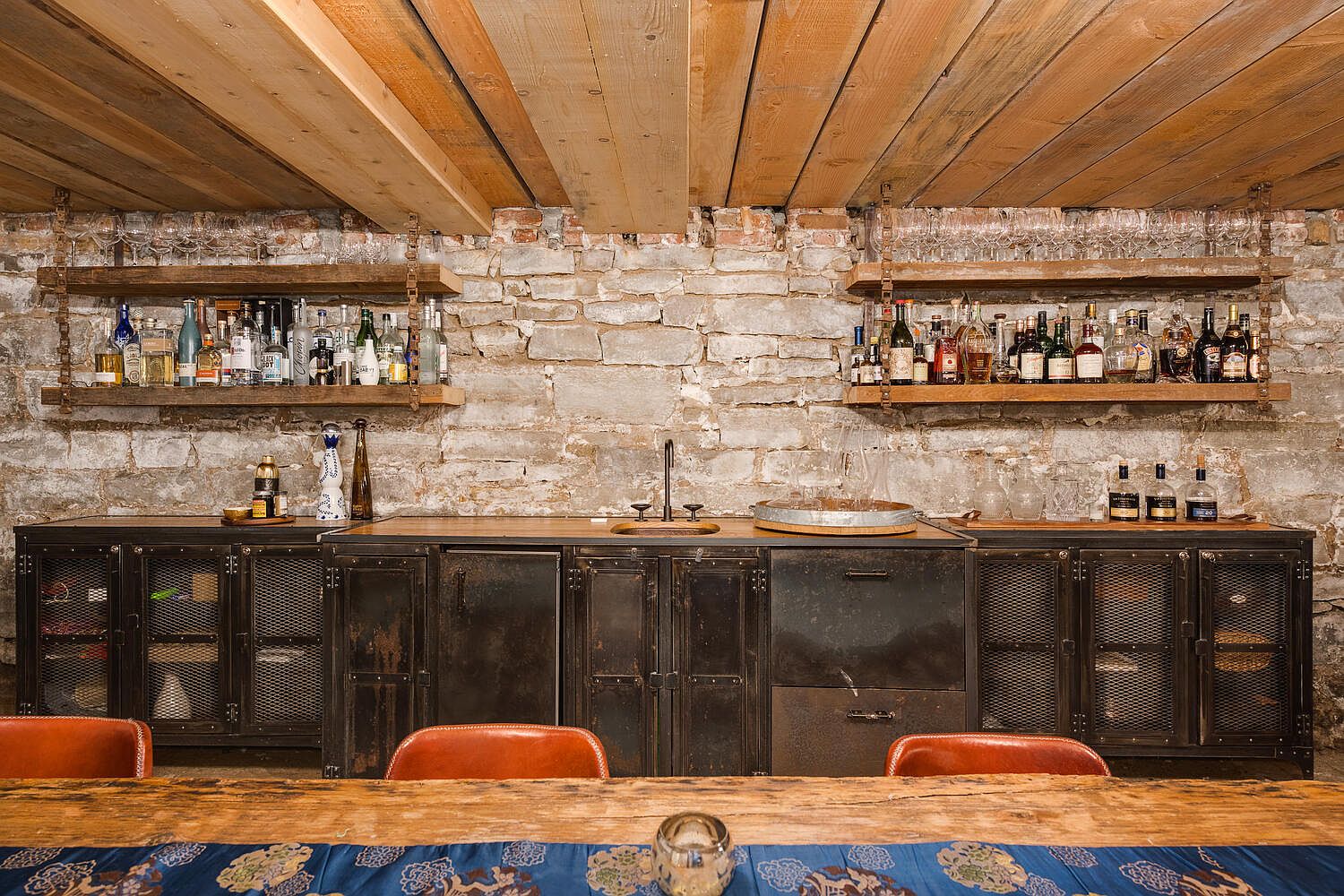 This image showcases a rustic wine cellar featuring exposed brick walls and a wooden plank ceiling. Two wooden shelves display an array of liquor bottles and glassware, while a metal cabinet with a wooden countertop houses a sink and additional storage. The scene is set with a wooden table and leather chairs, creating a cozy and inviting atmosphere.