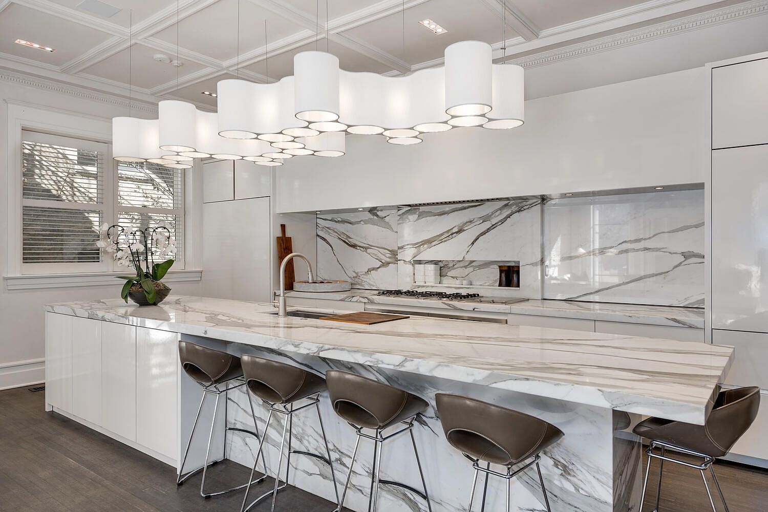 This is a bright, modern kitchen featuring a large marble island with seating, white cabinetry, and marble backsplash. The kitchen is illuminated by a unique, multi-tiered pendant lighting fixture. The overall impression is one of luxury and sophistication.