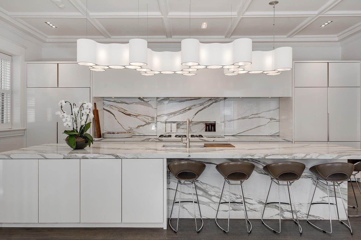 This is a modern kitchen featuring a large island with marble countertops and waterfall edges, complemented by sleek white cabinetry and a marble backsplash. A unique, multi-bulb pendant light fixture hangs above the island, and four bar stools are neatly arranged along one side. The kitchen exudes a luxurious and contemporary feel.