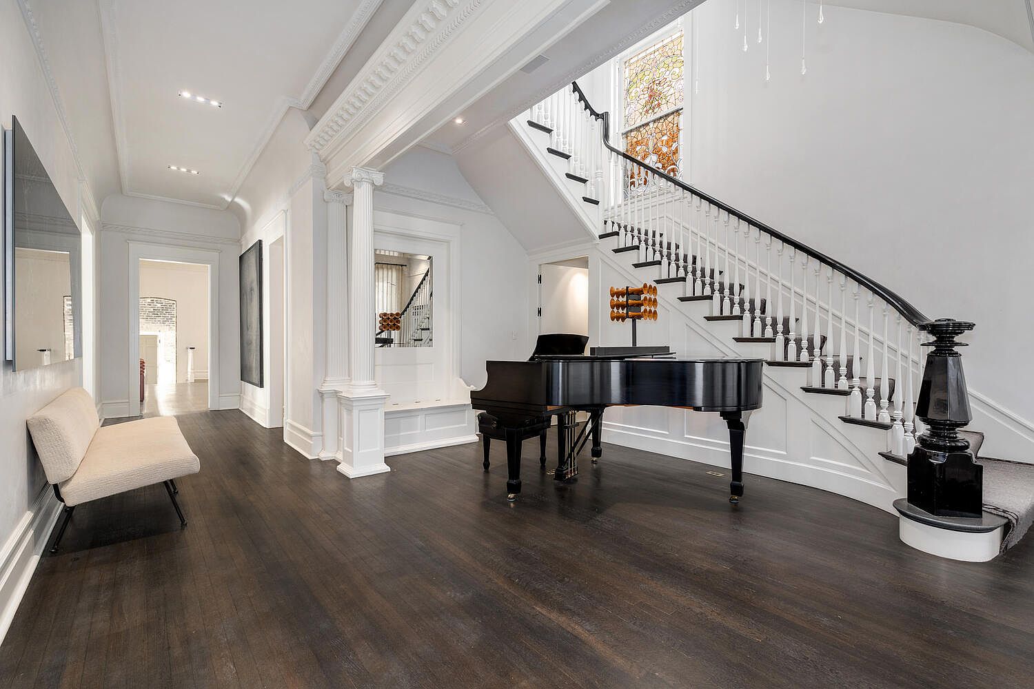 This grand foyer features dark hardwood floors, white walls, and a striking staircase with black accents. A black grand piano sits elegantly in the space, adding a touch of sophistication. The area is well-lit, highlighting the architectural details and creating a welcoming atmosphere.