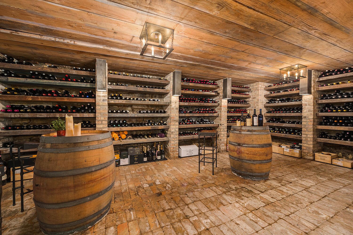 This is a well-stocked wine cellar featuring wooden shelves filled with numerous wine bottles. The room has a rustic aesthetic with brick walls, a brick floor, and a wooden plank ceiling. Two large wooden barrels serve as tables, adding to the cellar's charm and functionality.