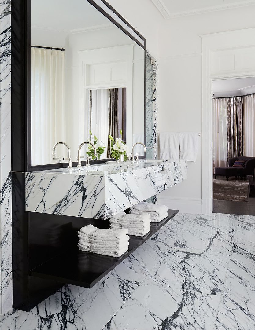 This is a luxurious primary bathroom featuring a double vanity with a marble countertop and backsplash. The black-framed mirror and dark wood shelf contrast with the white marble flooring and walls, creating a sophisticated and modern aesthetic. The open doorway reveals a glimpse of an adjacent living space, enhancing the sense of spaciousness.