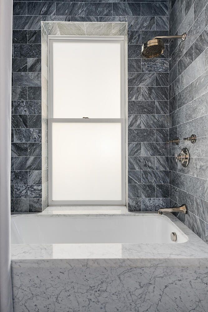 This is a well-lit primary bathroom featuring a white bathtub with a marble surround. The walls are tiled with gray stone, and a window provides natural light. The shower has a brass showerhead and fixtures, creating a luxurious and modern aesthetic.