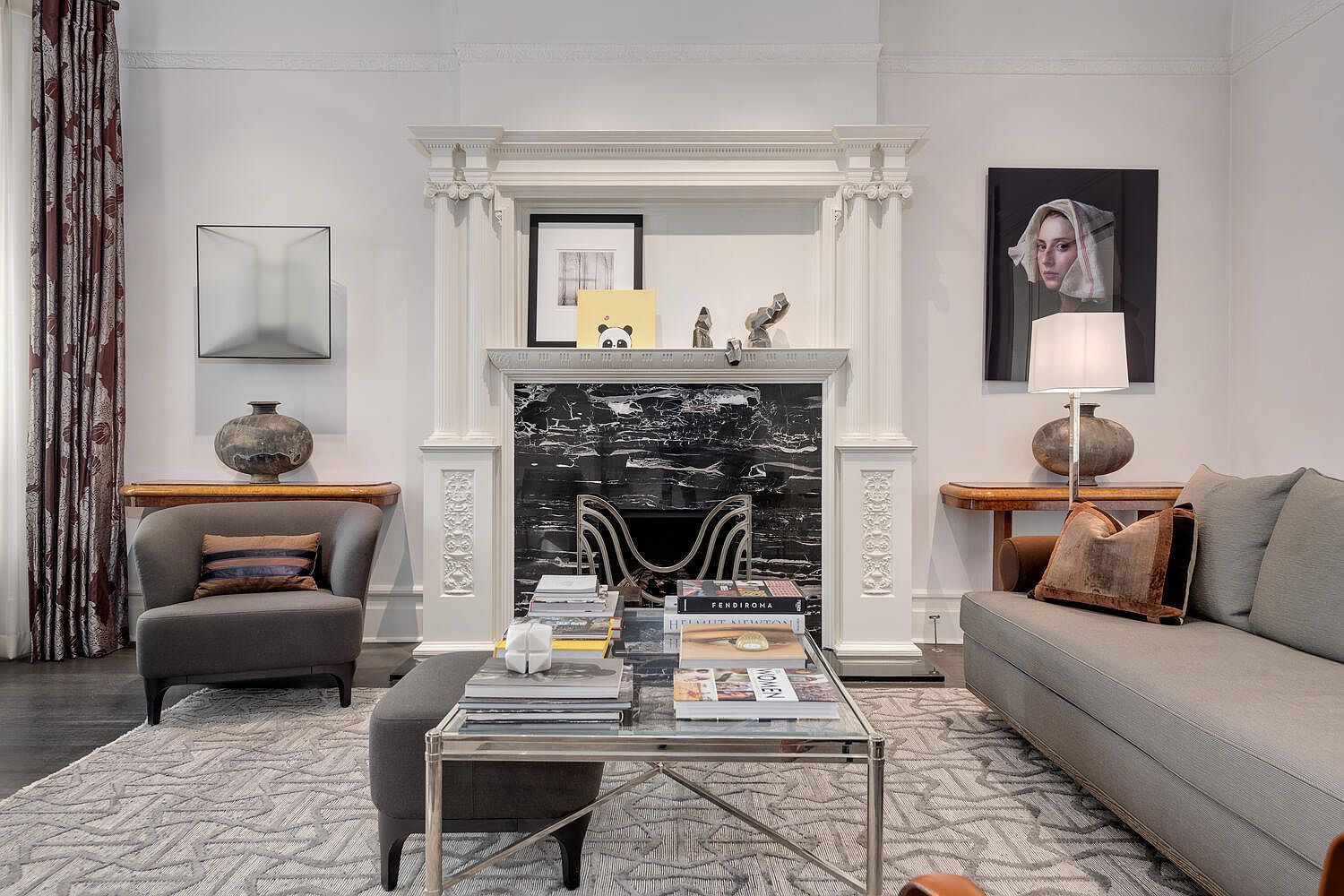 This is an interior shot of a living room featuring a fireplace with a black marble surround and a white mantel. The room is decorated with modern art and stylish furniture, including a gray sofa, armchair, and a glass-topped coffee table. The overall impression is elegant and sophisticated.
