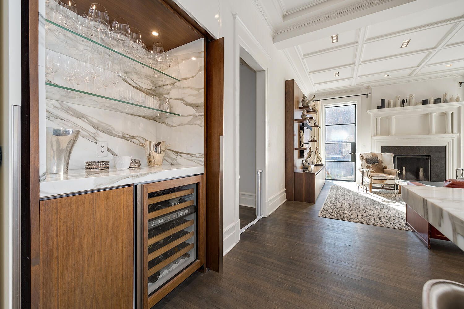 This interior shot showcases a sophisticated living space with a built-in bar area featuring marble accents and a wine cooler. The room transitions into a living area with a fireplace, elegant shelving, and a comfortable armchair, all enhanced by natural light streaming through a large window. The dark hardwood floors and coffered ceiling add to the room's luxurious feel.